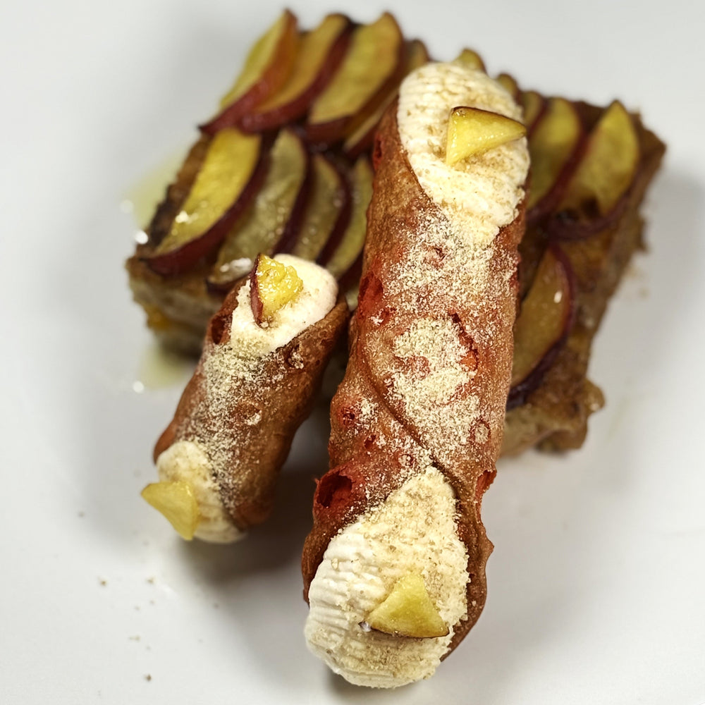 Add a playful fusion of Italian craft and Southern tradition with our Peach Cobbler Cannoli! Each crisp cannolo shell is filled with luscious ricotta cream infused with ripe, juicy peach purée and hints of warm cinnamon. Sprinkled atop, you'll find tender morsels of buttery pastry and spiced crumble, echoing the beloved cobbler’s classic flavours and textures. The ends are adorned with a sliver of fresh peaches.&nbsp; This cannolo delivers the nostalgia of a summer peach pie in an elegant, hand-held treat.&